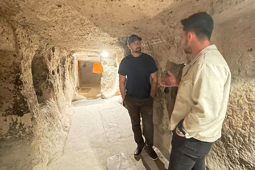 Kate’s husband listens to a guide while standing inside a narrow, dimly lit tunnel of an ancient underground city carved into the rock in Cappadocia, Turkey.