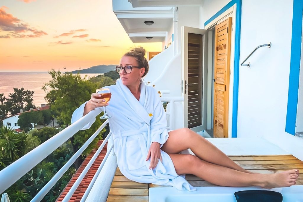 Kate relaxes in a white robe with a glass of wine on a balcony in Turkey, overlooking a coastal sunset framed by trees and hillside rooftops.