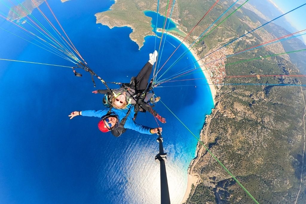 Kate paraglides high above the turquoise coastline of Ölüdeniz, Turkey, with her arms outstretched and a wide smile, soaring over the famous Blue Lagoon.