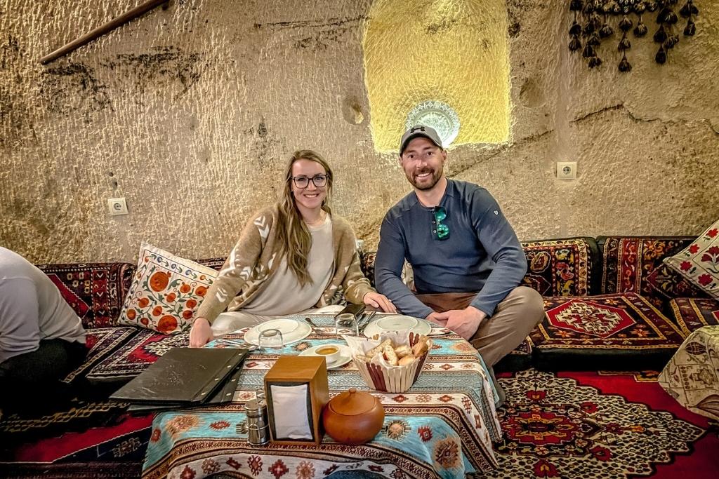 Kate and her husband sit together on vibrant, patterned cushions at a traditional Turkish restaurant carved into a stone wall, smiling with tea and bread in front of them.