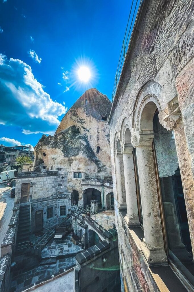 Bright sunlight shines over the rocky facade of a cave hotel in Cappadocia, with arched stone windows and terraces built into the natural rock formations.