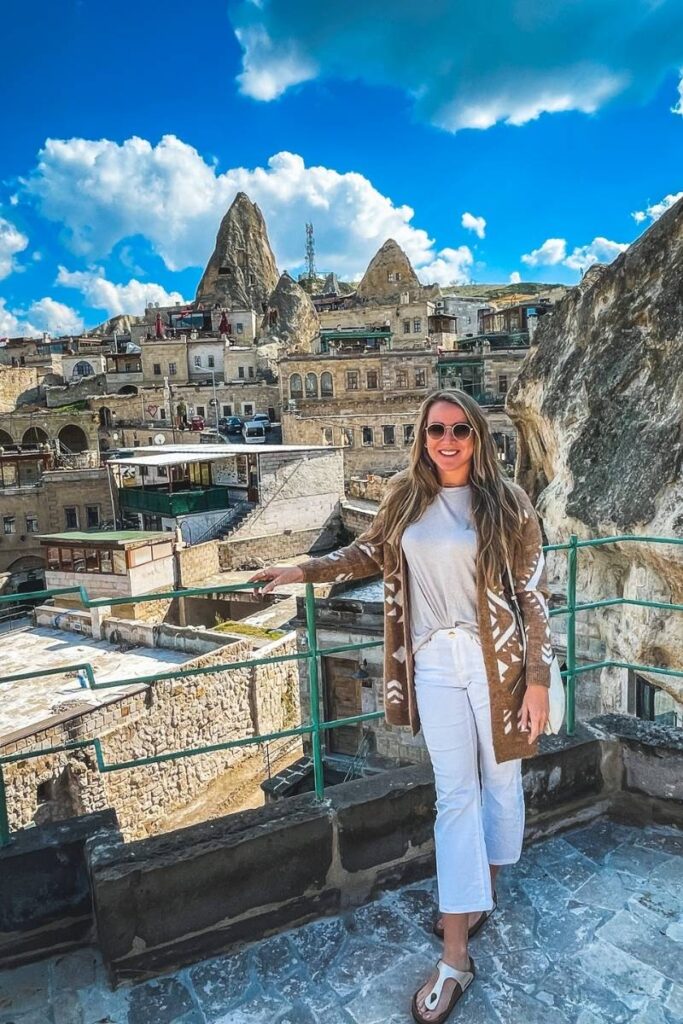 Kate smiles on a rooftop terrace in Cappadocia, with a stunning view of stone buildings and rocky peaks rising under a bright blue sky.