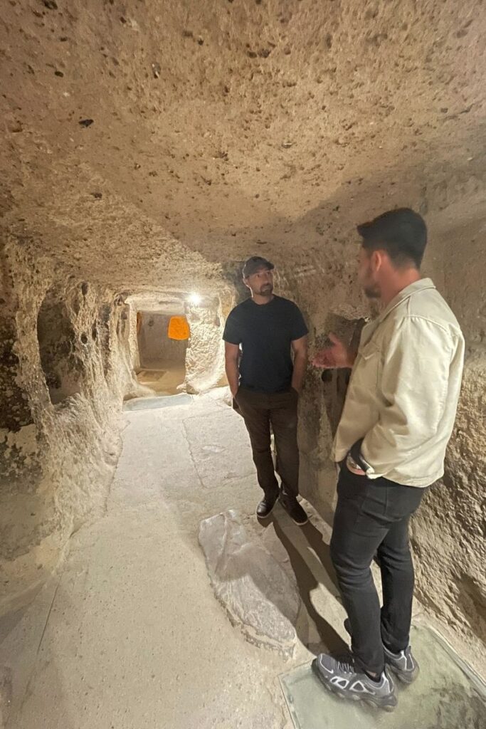 Two men talk inside a softly lit passage of Cappadocia’s underground city, surrounded by carved stone walls and narrow hallways.