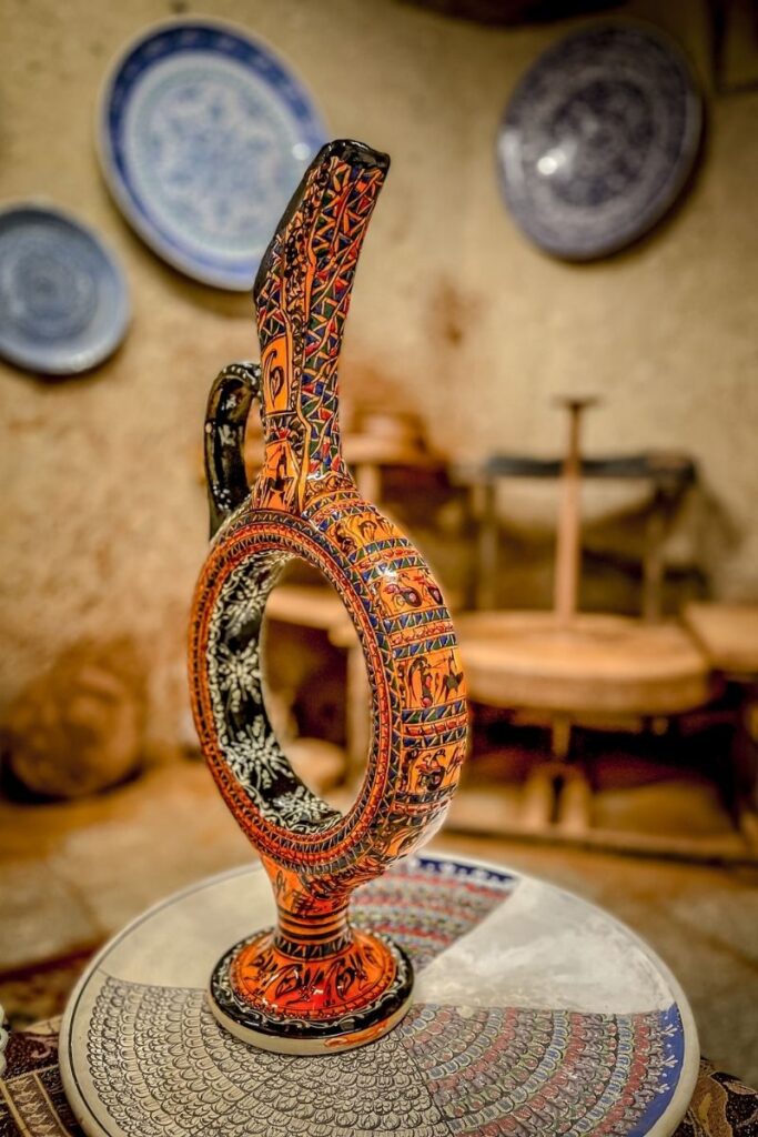 A vibrant, hand-painted Hittite-style wine jug with an open circular center sits on a decorated ceramic table in a pottery studio in Cappadocia.
