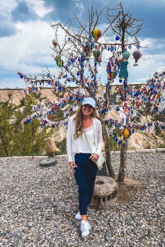 Kate poses in front of a tree decorated with evil eye charms, ribbons, and hot air balloon ornaments in Cappadocia, with rocky hills in the background.