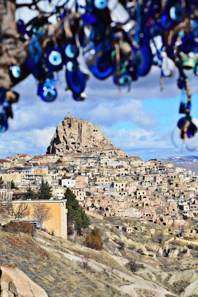 A scenic view of Uçhisar Castle and the surrounding cave dwellings in Cappadocia, framed by hanging evil eye charms in the foreground.