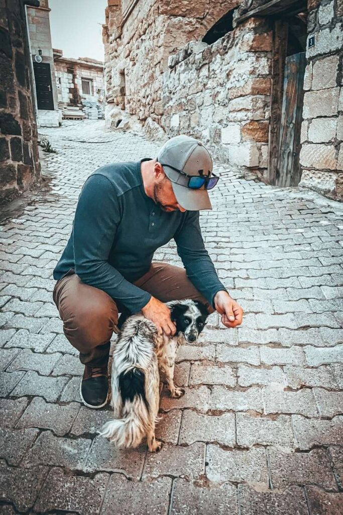 Kate’s husband kneels to gently pet a small, scruffy dog on a quiet cobblestone street lined with old stone buildings in Cappadocia.