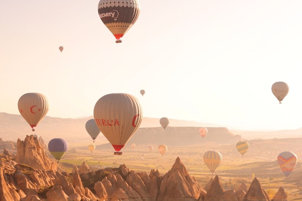 Dozens of hot air balloons drift above Cappadocia’s jagged rock formations at sunrise, with soft golden light illuminating the valley below.