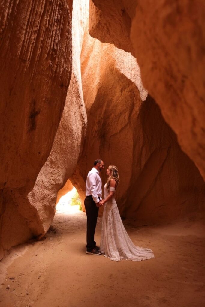 Kate and her husband stand hand in hand inside a narrow, sunlit cave in Cappadocia, framed by towering terracotta-colored rock walls.