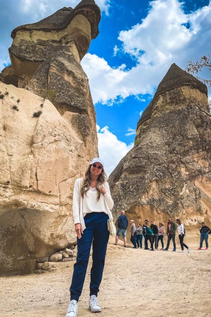 Kate smiles in front of Cappadocia’s iconic fairy chimneys under a bright blue sky, with other tourists exploring the rock formations in the background.