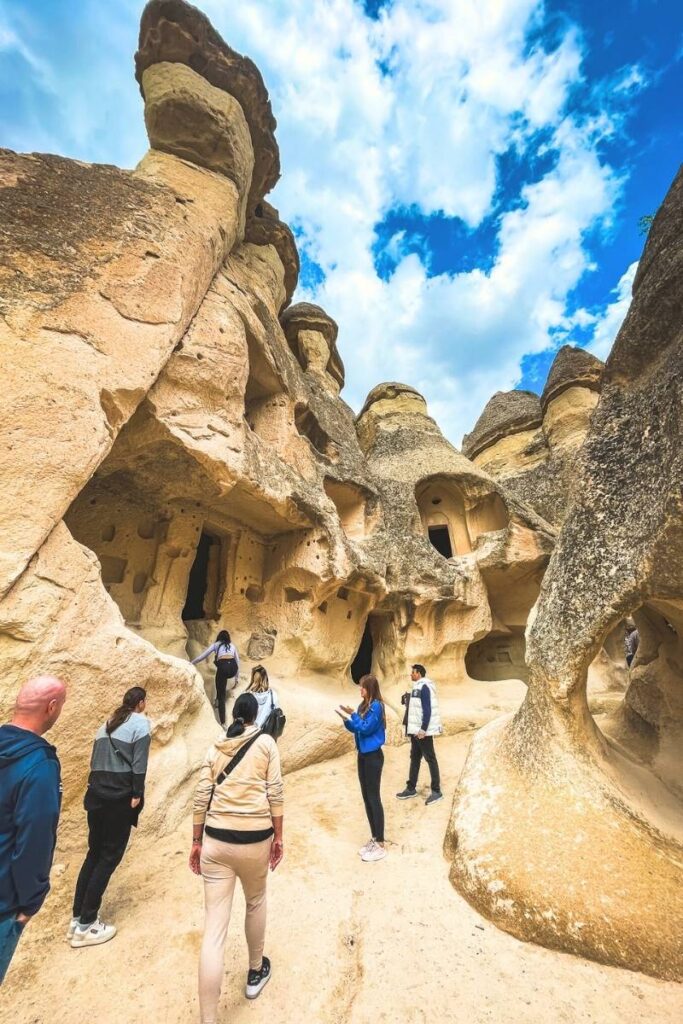 Visitors explore the ancient cave dwellings carved into Cappadocia’s fairy chimneys, surrounded by textured rock formations and a vibrant blue sky overhead.
