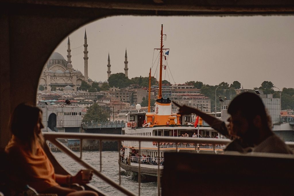 Framed by a ferry window, Kate holds a cup while gazing toward the skyline of Istanbul, where the Suleymaniye Mosque and a docked ferry stand out against the hazy afternoon light.