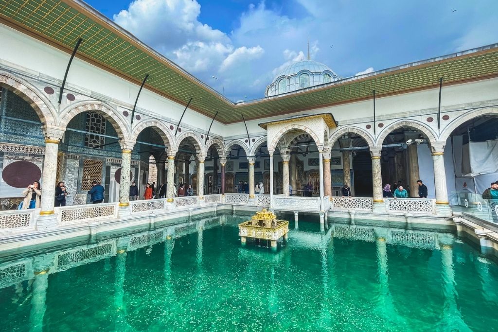 An ornate courtyard pool reflects the arches and columns of Topkapı Palace’s Harem section, with visitors strolling under golden-trimmed ceilings and intricate tilework beneath a blue sky.