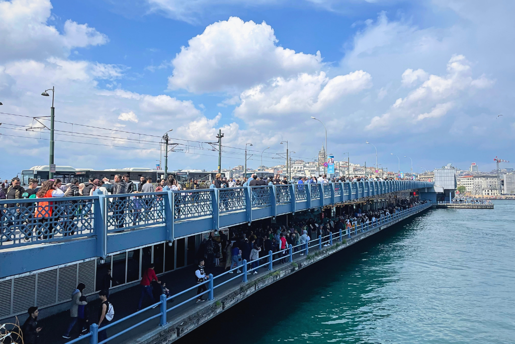 Locals line the upper level of Galata Bridge with fishing rods while people dine and stroll along the waterfront restaurants below, set against Istanbul’s historic skyline.