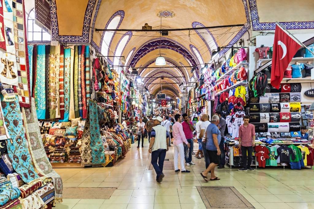 Shoppers browse the vibrant aisles of Istanbul’s Grand Bazaar, lined with colorful textiles, Turkish flags, souvenirs, and arched ceilings adorned with traditional tilework.