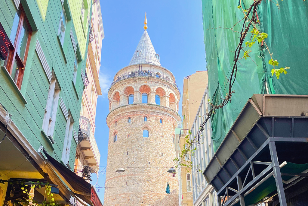 The iconic stone Galata Tower rises between narrow buildings in Istanbul, its cylindrical structure and pointed roof glowing under a bright blue sky.