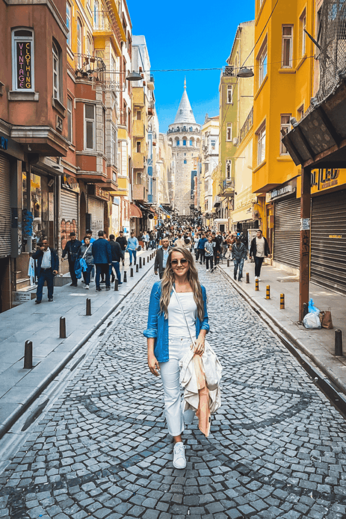 Kate stands smiling on a cobblestone street lined with colorful buildings, with the Galata Tower rising above a bustling crowd in the background under a clear blue sky.