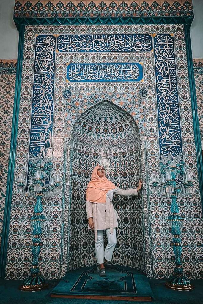 Kate stands in front of an ornately tiled mihrab inside a mosque, surrounded by intricate floral and geometric Iznik tilework, Arabic calligraphy, and decorative turquoise lanterns.