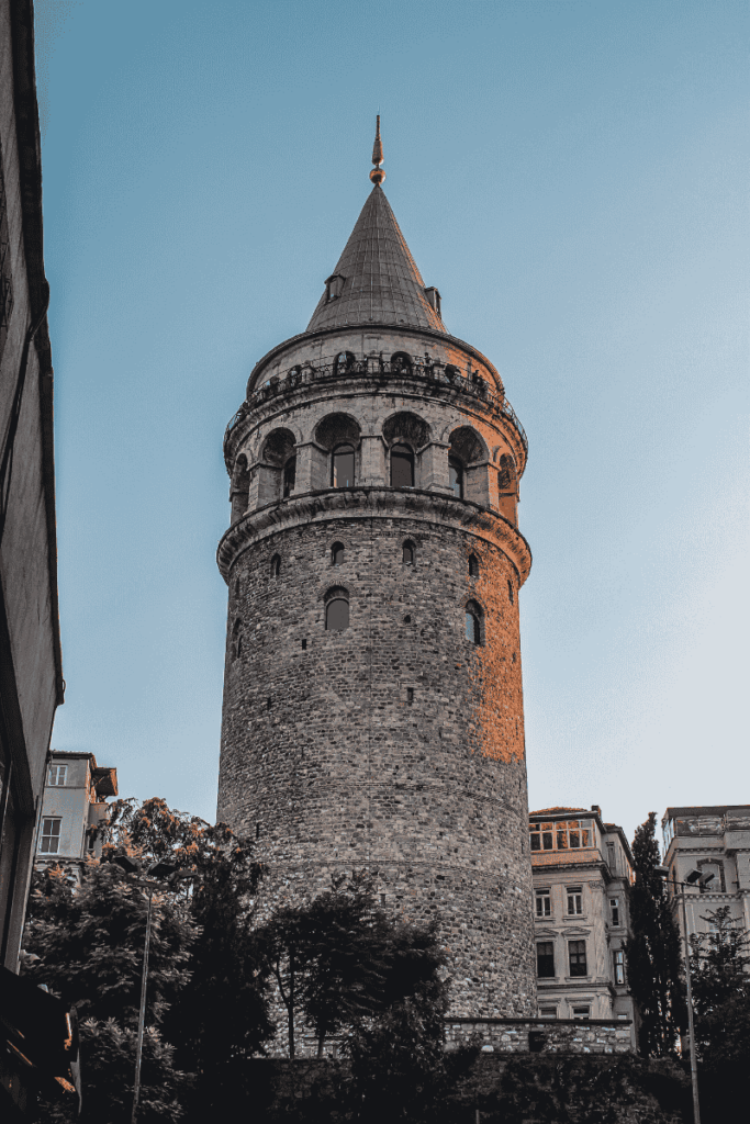 The Galata Tower stands tall with its medieval stone walls and conical roof bathed in golden evening light, framed by nearby buildings and trees against a clear sky.
