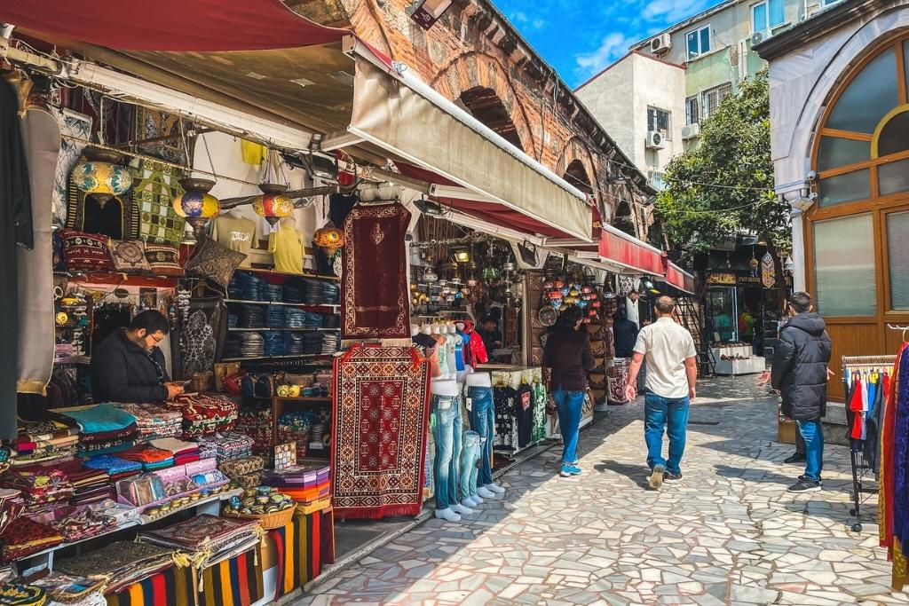 Shoppers stroll through an outdoor section of Istanbul’s Grand Bazaar, passing stalls filled with colorful textiles, mosaic lanterns, rugs, and traditional Turkish goods under awnings and open sky.