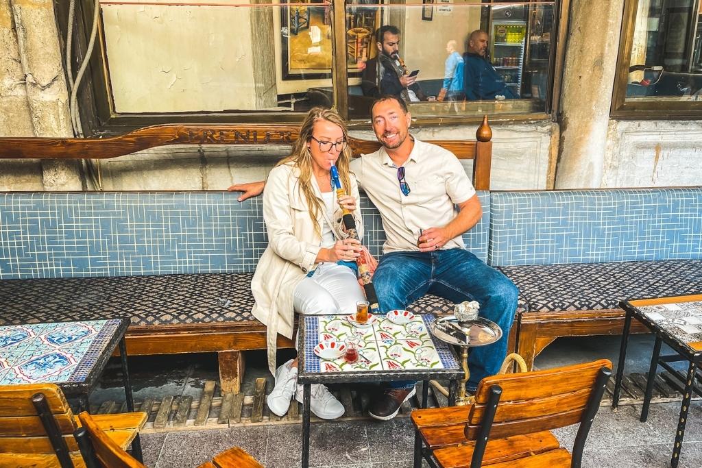 Kate and her husband relax at a traditional Istanbul café, sipping Turkish tea and sharing a hookah while seated on a tiled bench surrounded by colorful mosaic-topped tables.