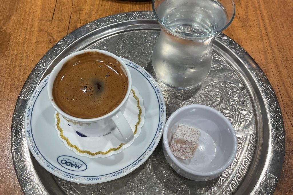 A traditional serving of Turkish coffee sits on an ornate silver tray, accompanied by a glass of water and a small bowl holding a single piece of powdered Turkish delight.