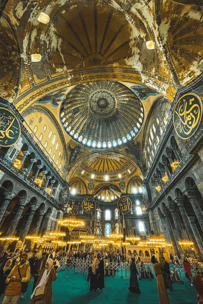 Visitors stand beneath Hagia Sophia’s massive domed ceiling and glowing chandeliers, surrounded by grand arches, Arabic calligraphy medallions, and weathered historic walls.
