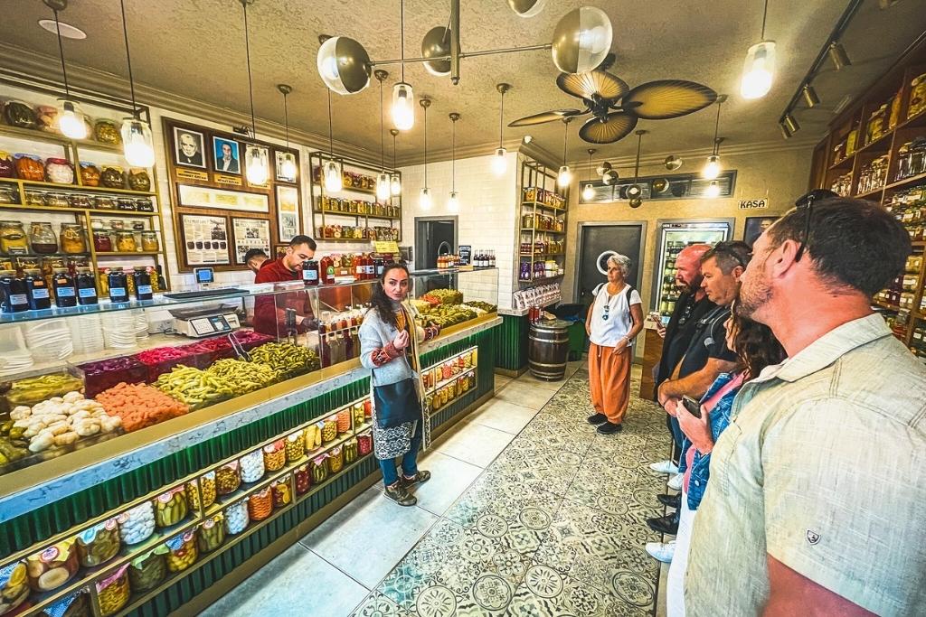 A small group listens to a woman explaining local produce inside a colorful Turkish pickle shop, with shelves and counters overflowing with jars of preserved vegetables and spices.