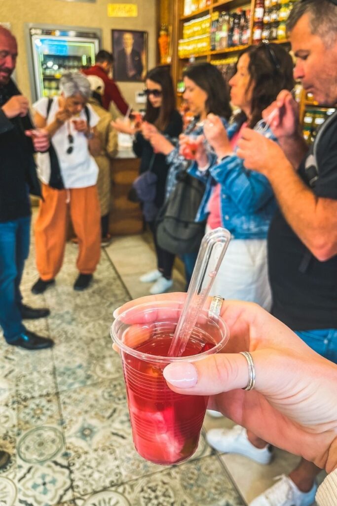 Close-up of a plastic cup filled with red pickle juice and vegetables, held up during a tasting session in a vibrant pickle shop as a group of tourists sample and react in the background.