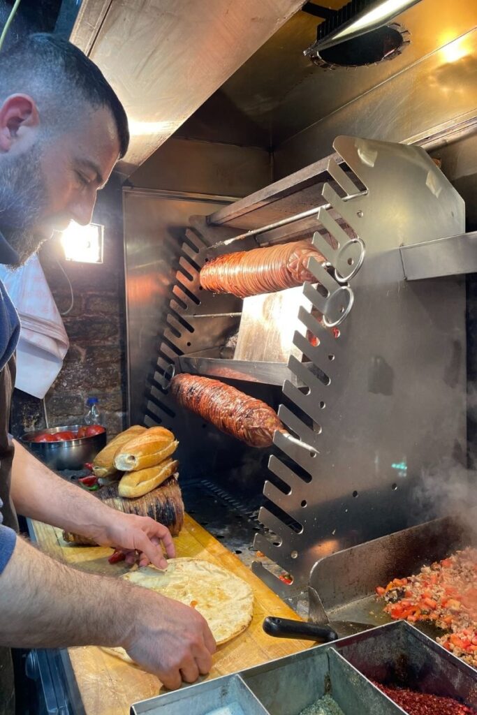 A street food vendor in Istanbul prepares a sandwich using kokoreç, a traditional Turkish dish made from seasoned lamb intestines grilled on horizontal skewers, with fresh bread and chopped tomato mixture nearby.