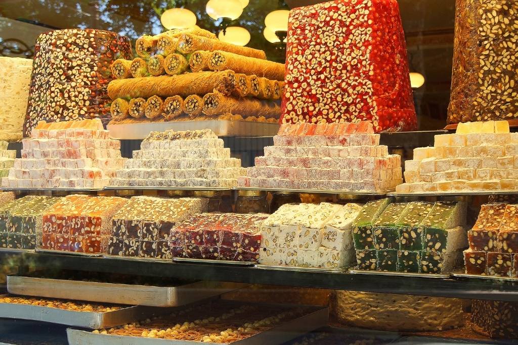 A vibrant shop window stacked with colorful Turkish delights and nut-studded nougats, including pyramid piles dusted in powdered sugar, rolled pistachio treats, and towering blocks of nut-packed confections.