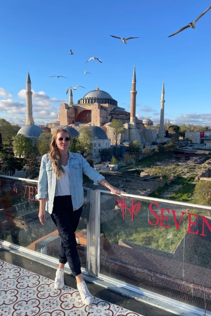 Kate poses on a rooftop terrace with the iconic Hagia Sophia in Istanbul as a breathtaking backdrop, surrounded by blue skies and seagulls gliding above the historic skyline.
