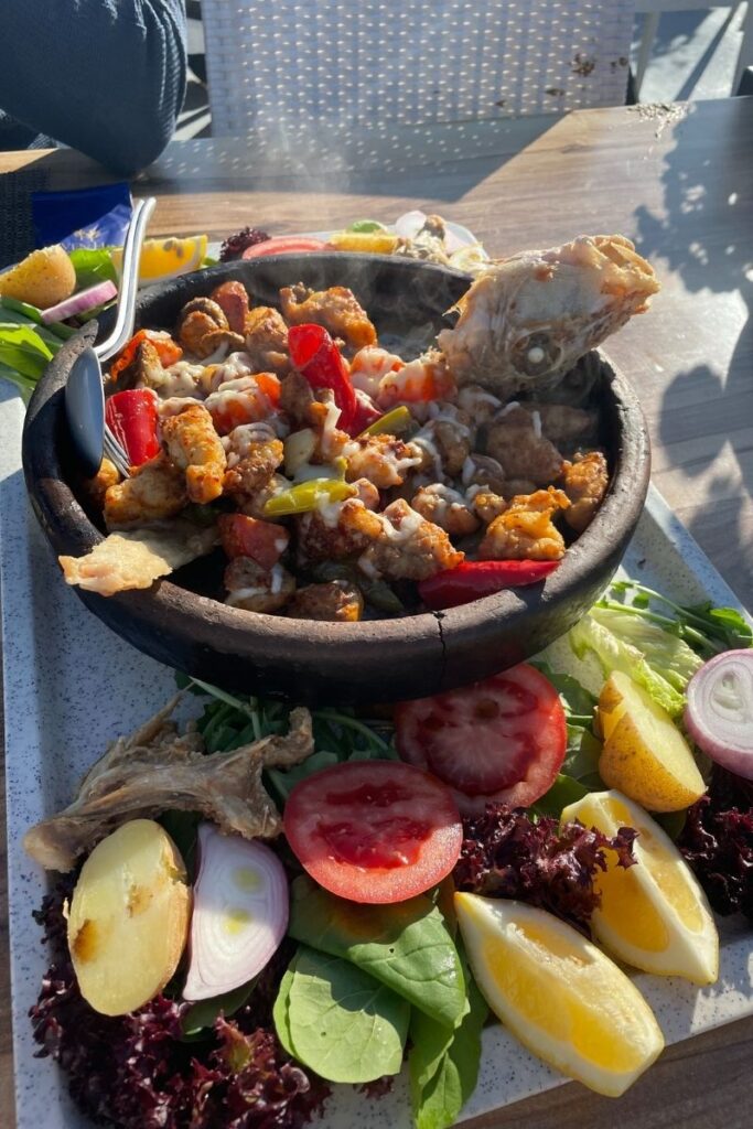 A sizzling hot clay pot of Turkish-style sautéed fish and vegetables, topped with melted cheese and served with a colorful side of fresh salad including tomatoes, onions, lemon wedges, and leafy greens.