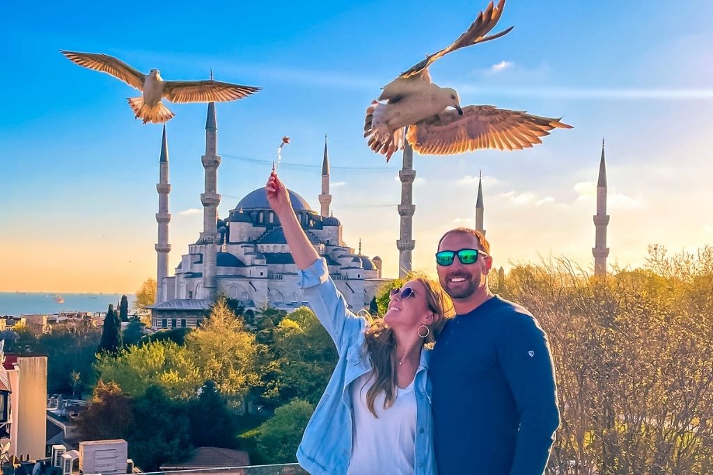 Kate and her husband pose joyfully on a rooftop at sunset with the iconic Blue Mosque in the background, as seagulls swoop in mid-air to snatch food from the woman's outstretched hand, creating a lively and magical moment.