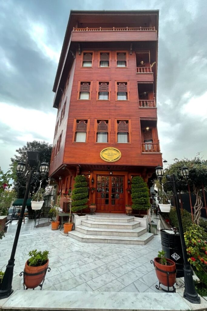 Front view of the Hotel Valide Sultan Konagi, a traditional wooden building in Istanbul with ornate windows, potted plants flanking the marble steps, and a charming, historic Ottoman architectural style under a moody sky.