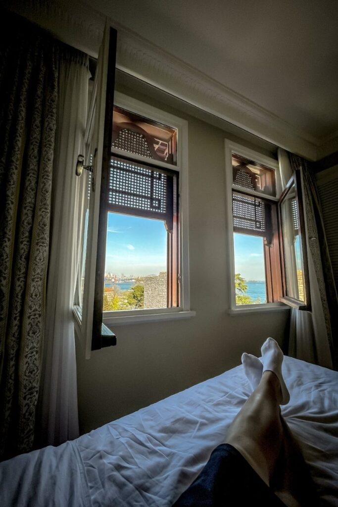 A relaxed view from a hotel bed looking out through two open wooden windows at the sparkling Bosphorus and Istanbul skyline under a clear blue sky, with Kate's legs stretched out in the foreground.