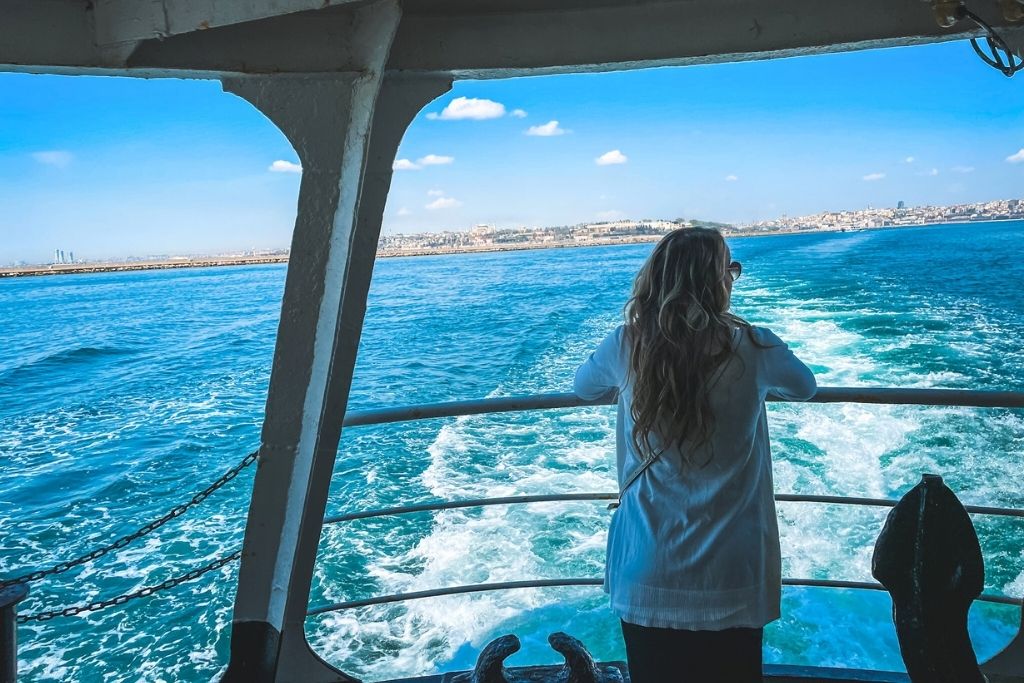 Kate stands at the back of a ferry gazing out over the vivid blue waters of the Bosphorus Strait, with the wake trailing behind the boat and the skyline of Istanbul stretching along the horizon under a sunny sky.