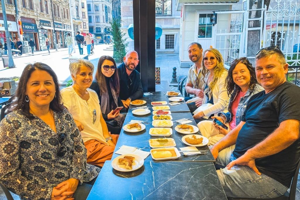 A group of nine people smile around a long black table at a street-side café in Istanbul, enjoying a Turkish breakfast with an array of small dishes including olives, cheeses, spreads, and breads laid out in front of them.