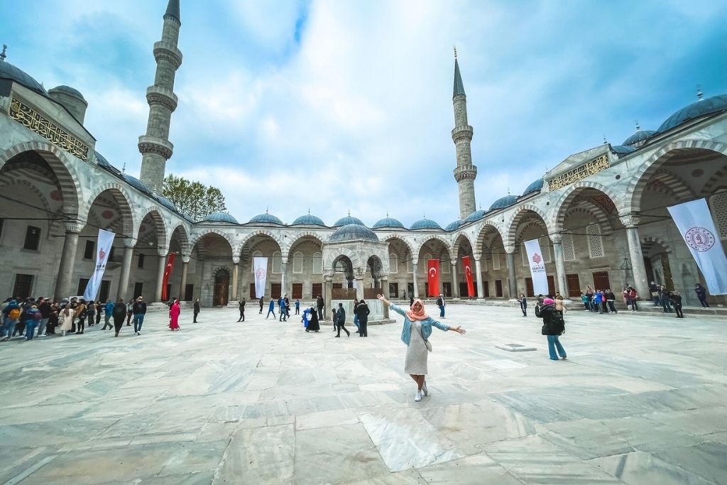 Kate stands with arms outstretched in the grand courtyard of the Blue Mosque (Sultan Ahmed Mosque) in Istanbul, surrounded by arched colonnades, domes, and towering minarets under a dramatic cloudy sky.