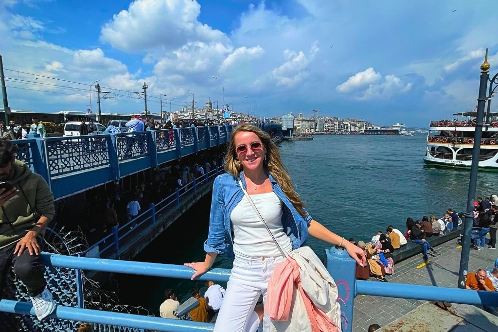Kate smiles in the sunshine while leaning against a blue railing by the water, with Galata Bridge, ferry traffic, and the Istanbul cityscape stretching out behind her.