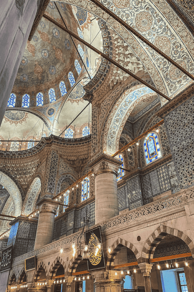 An angled view of the Blue Mosque’s interior reveals intricate floral and geometric tilework, ornate arches, stained glass windows, and a soaring dome illuminated by soft daylight.