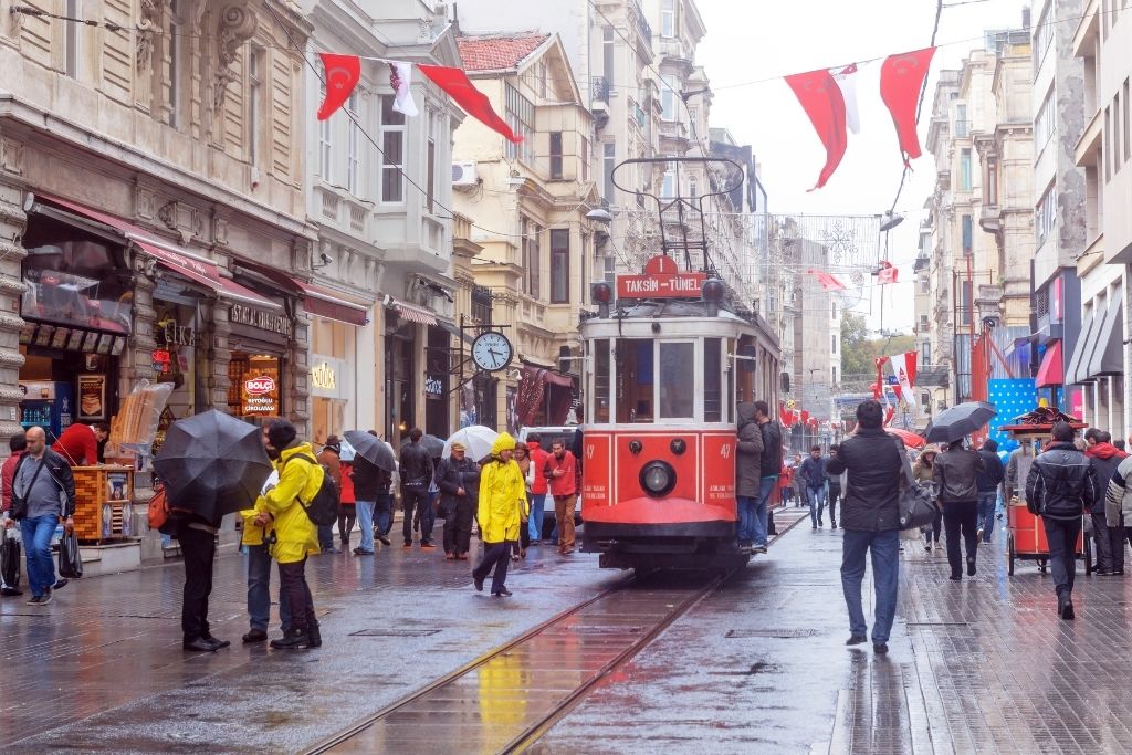 A red vintage tram marked “Taksim - Tünel” travels down a rainy Istiklal Street in Istanbul, as people with umbrellas and Turkish flags overhead add color to the bustling pedestrian avenue.