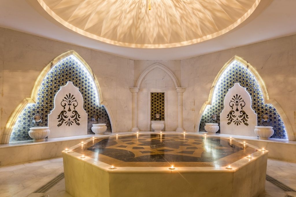 A luxurious Turkish hammam features a central marble platform surrounded by ornate tiled alcoves, soft lighting, and decorative fountains, creating a serene and elegant atmosphere.