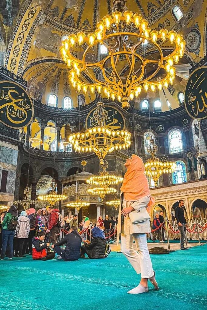 Kate, barefoot and draped in a peach headscarf, gazes upward in awe beneath golden chandeliers inside the vibrant, domed interior of Hagia Sophia.