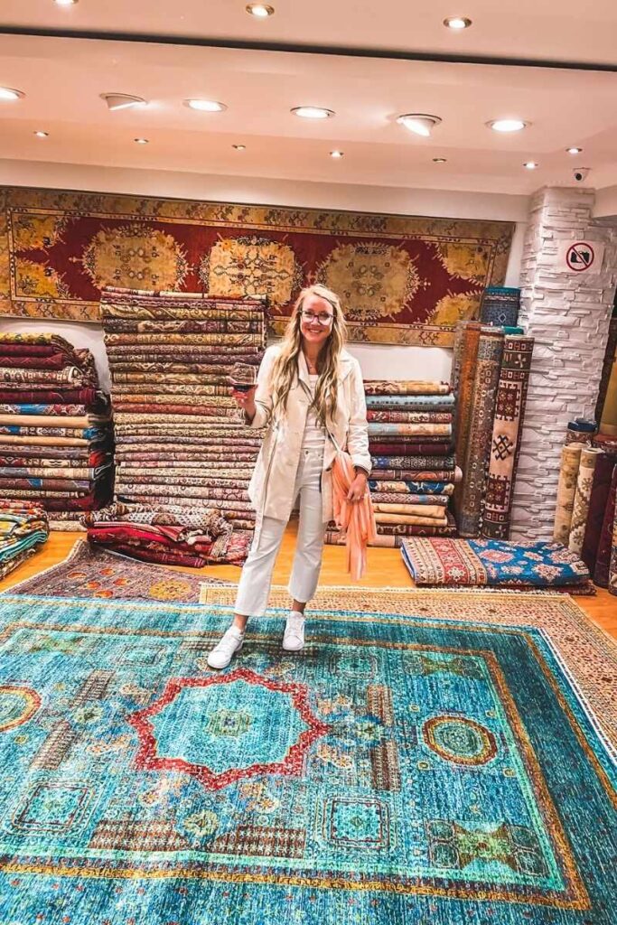 Kate stands barefoot on a turquoise Turkish rug, holding a glass of tea and smiling inside a vibrant carpet shop lined with stacks of intricately patterned carpets and kilims.