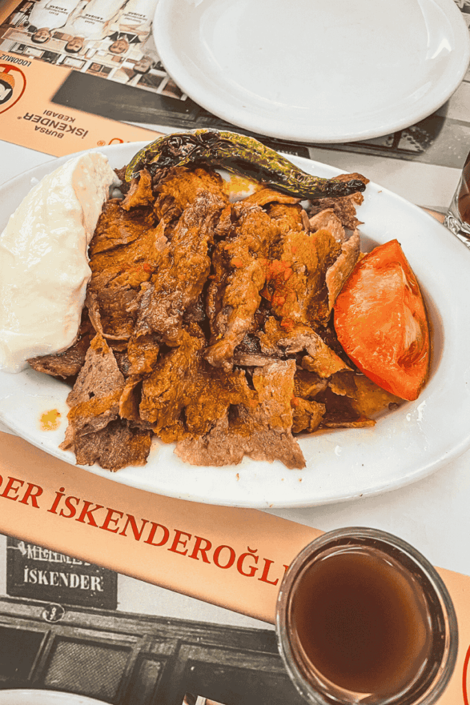 A plate of classic İskender kebab topped with thinly sliced lamb, tomato sauce, grilled pepper, roasted tomato, and a scoop of yogurt, served on a branded placemat with a glass of şıra.