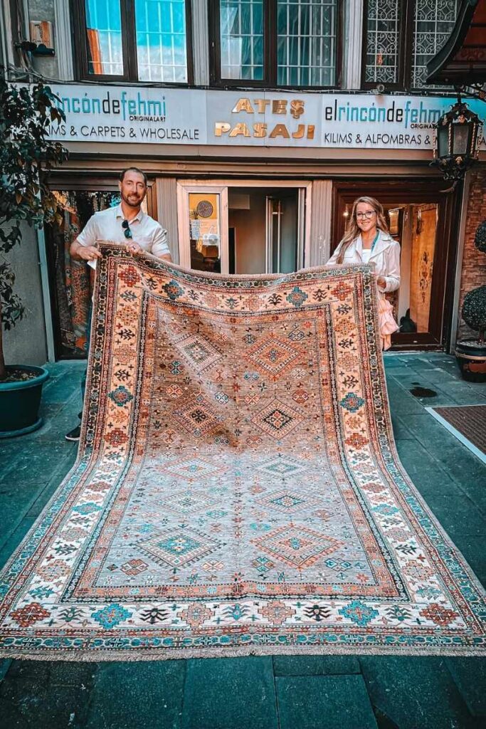 Kate and a man hold up a large, intricately patterned Turkish rug outside a carpet shop in Istanbul, with geometric motifs in earthy tones and teal accents on display.