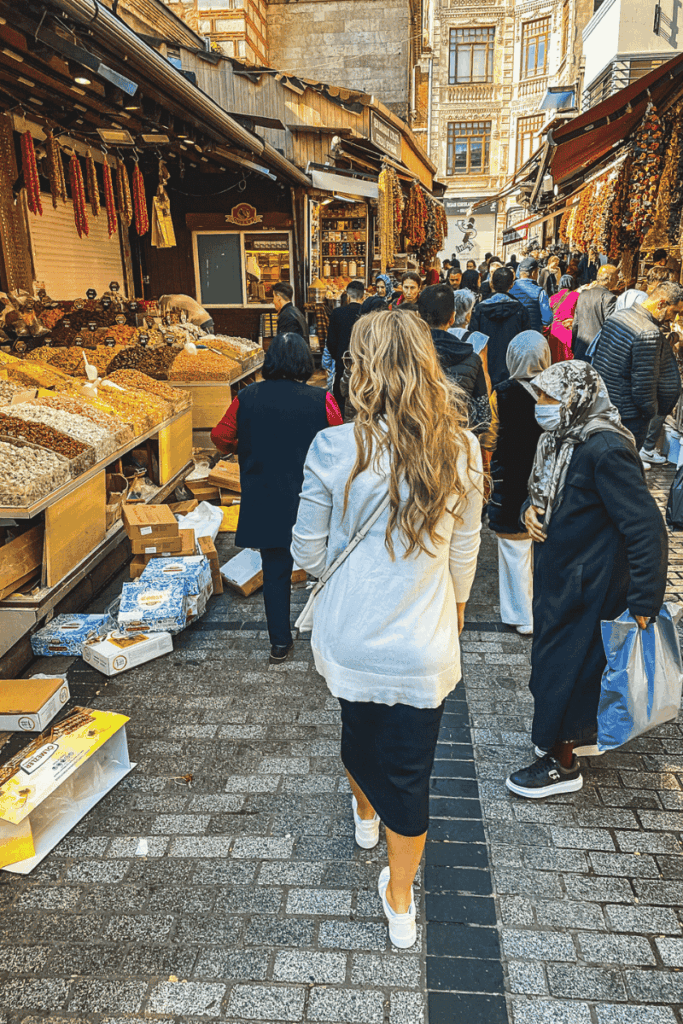 Kate walks through a bustling open-air spice and nut market in Istanbul, surrounded by hanging dried peppers, local shoppers, and overflowing bins of dried goods.