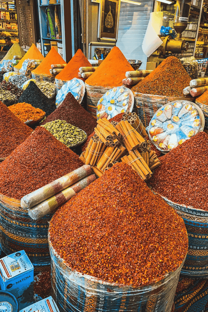Vibrant pyramids of loose spices—like chili flakes, turmeric, sumac, and cardamom—fill baskets at an Istanbul spice market, accented by bundles of cinnamon sticks and wrapped spice tubes.