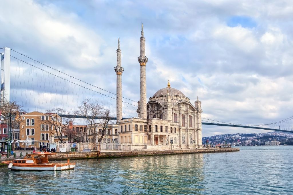 The ornate Ortaköy Mosque sits gracefully on the Bosphorus waterfront in Istanbul, framed by the Bosphorus Bridge and a wooden boat floating in the calm, reflective water.